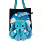 A blue Dysautonomia Rabbit plush sits in front of a black tote bag with a blue umbrella and cloud design.