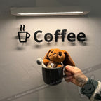 A Latte Lapin plushie bunny sits in a coffee mug, held up before a wall sign that reads Coffee with a cup icon.