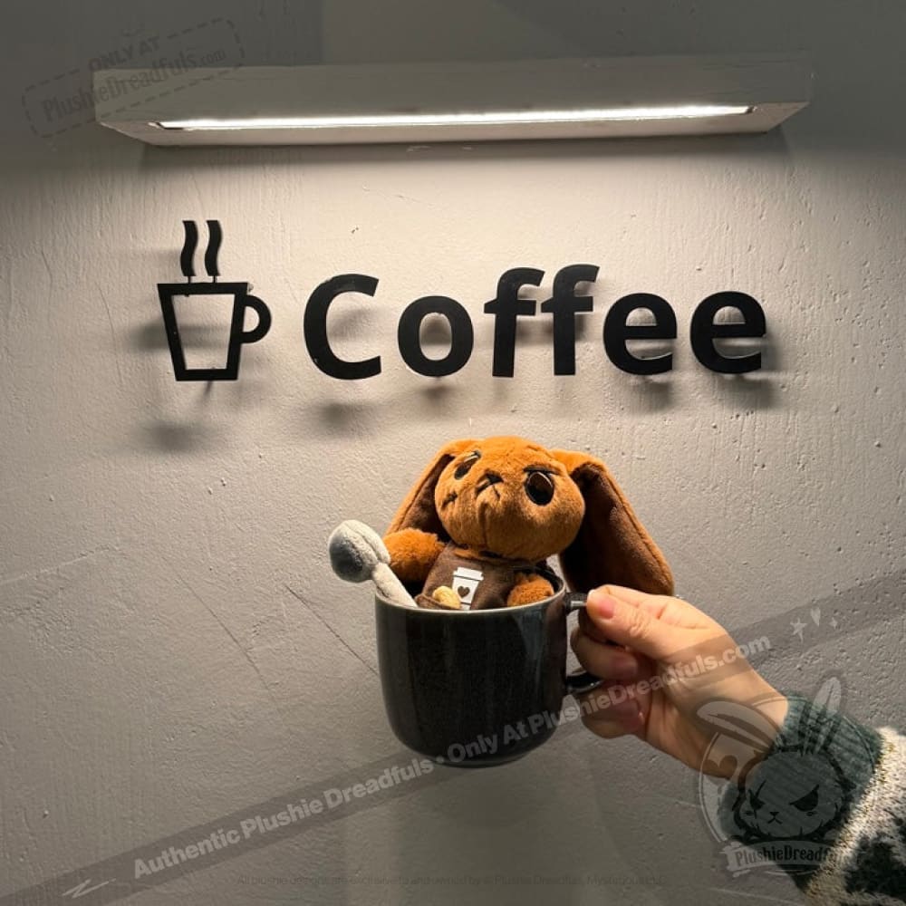 A Latte Lapin plushie bunny sits in a coffee mug, held up before a wall sign that reads Coffee with a cup icon.