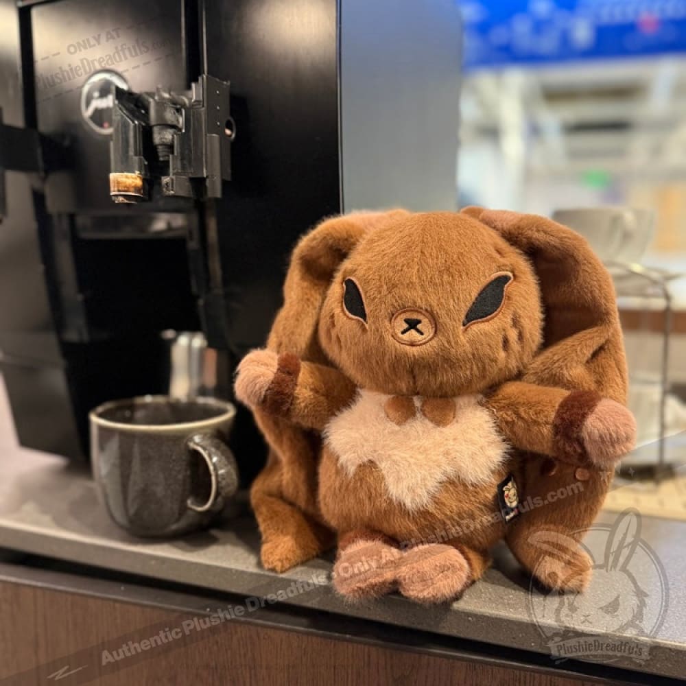 A Latte Lapin plushie sits next to a black coffee machine and mug on a kitchen countertop, ready for coffee time.