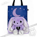 A purple plush bunny with a broken heart and semicolon symbol sits by a blue tote with crescent moon and stars.