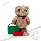 A brown Plushie Dreadfuls cat with broken heart patch, green bag, and red toy sits on white—find it at plushiedreadfuls.com.