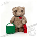 A Plushie Dreadfuls bear with a heart patch holds a green bag and red item, from plushiedreadfuls.com.