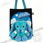 A blue Dysautonomia Rabbit plush sits in front of a black tote bag with a blue umbrella and cloud design.