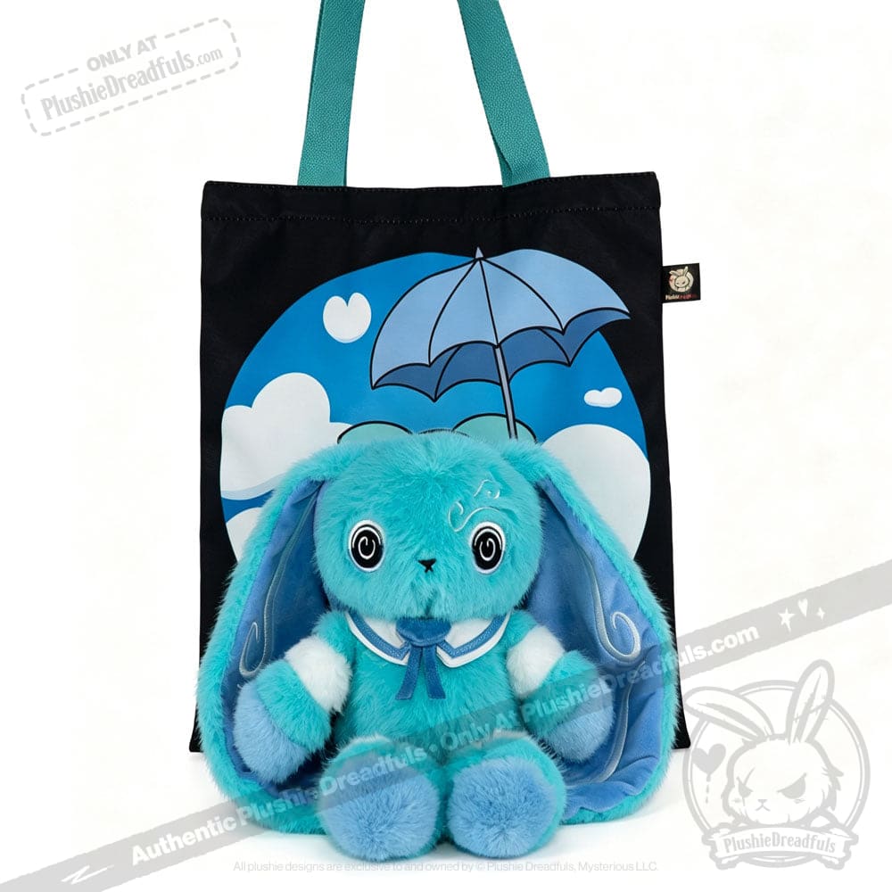 A blue Dysautonomia Rabbit plush sits in front of a black tote bag with a blue umbrella and cloud design.