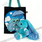 Blue plush Dysautonomia Rabbit sits beside a black tote bag featuring the bunny with an umbrella on a blue background.
