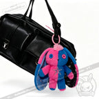 A black handbag with a Gemini Rabbit plushie keychain in pink and blue hanging from one of its handles.