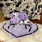 Two purple plush bunnies sit on a purple goth floor rug, bows on top, with a black and white forest tapestry behind.