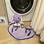 A purple stuffed bunny sits on a purple goth floor rug shaped like a heart in front of a washing machine.