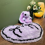 Plush bunny and flowers sit behind a purple goth floor rug, heart-shaped with black ribbons, on a wooden table.