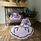 Two plush bunnies and a purple goth floor rug sit near potted plants on a wooden floor and shelf.