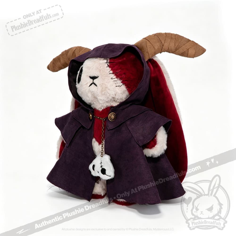 A Plushie Dreadfuls bunny with a horned cloak and red cape holds a skull charm, set against a white background.