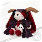 A Plushie Dreadfuls bear with ram horns, a red cape, and purple plushie cloak cuddles a matching mini plush in its lap.