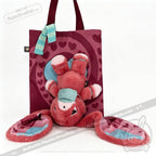 A pink awareness rabbit hangs upside down from a maroon heart tote bag, wrapped in a blue-striped scarf.