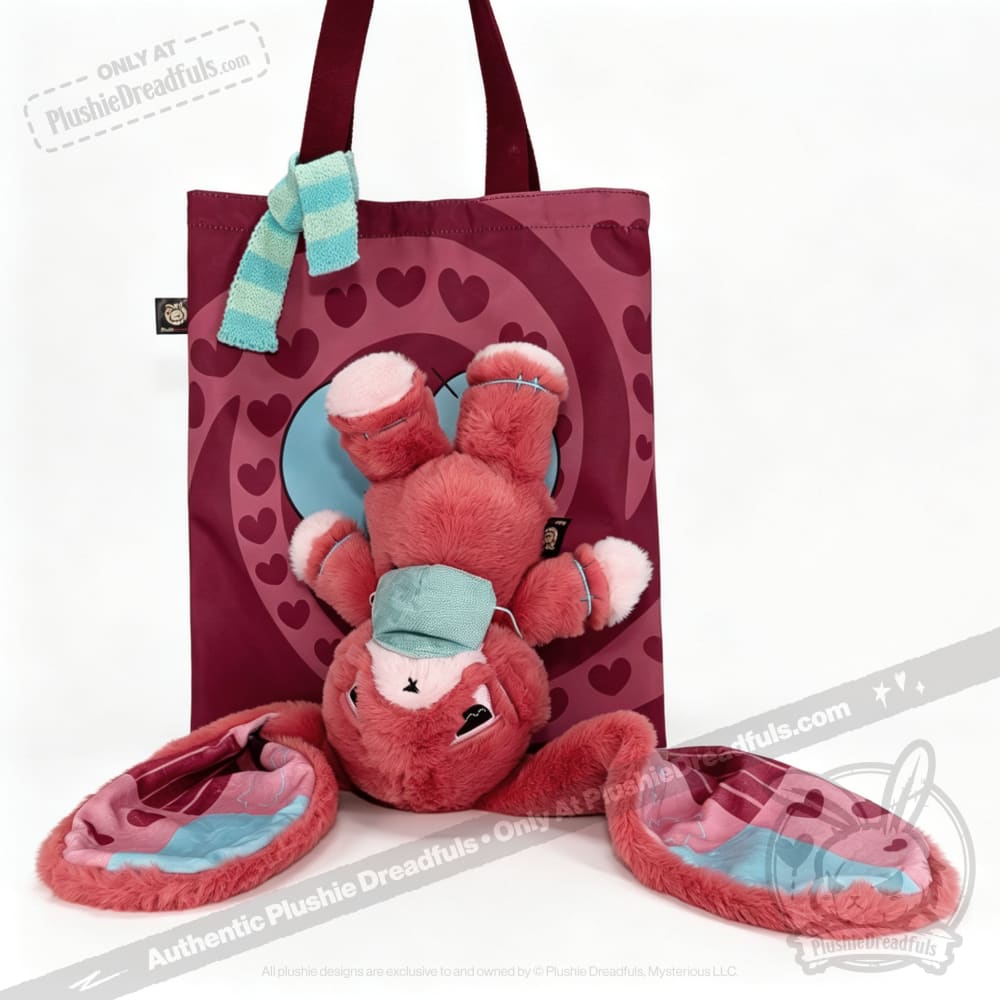 A pink awareness rabbit hangs upside down from a maroon heart tote bag, wrapped in a blue-striped scarf.