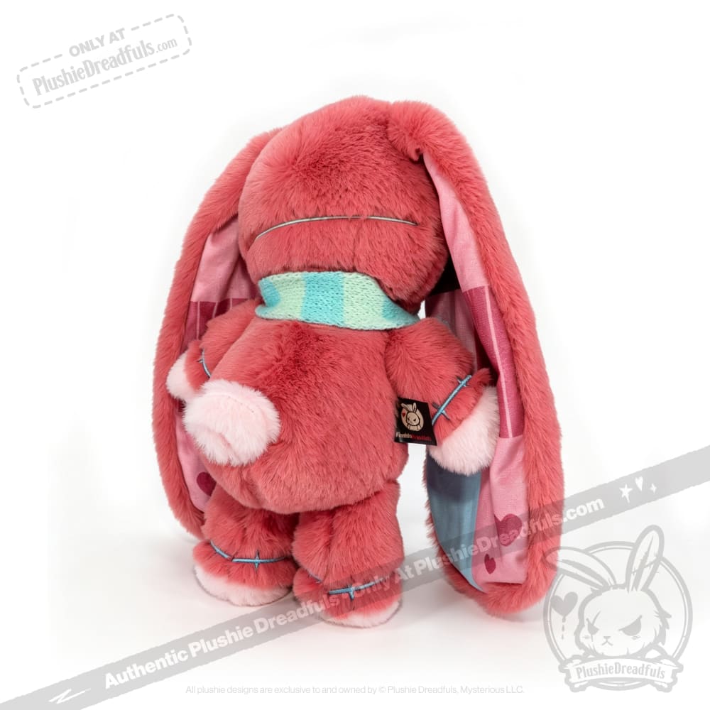 Plush pink awareness rabbit with floppy ears, blue scarf, and heart lining, facing away on a white background.