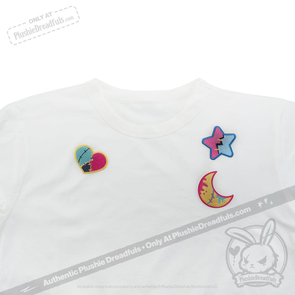 White T-shirt with three colorful iron on patches—a heart, star, and crescent moon—on the upper left side.