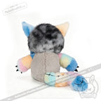 Presenting the Victorian McGee's Autism Acceptance Cheshire Cat - Plush Stuffed Animal: this whimsical plush features a gray furry head, pastel rainbow ears, arms, and tail, a beige body, and tiny claws. Inspired by the Alice in Wonderland plush style, it is displayed facing away to reveal its back and a stitched smile on its tail—a delightful tribute to autism acceptance.