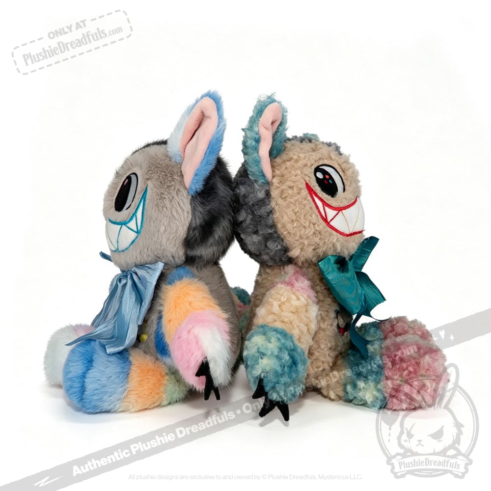 Two vibrant, furry plush toys sit back to back. Each features oversized grinning mouths with sharp red teeth, large round eyes, and blue bows—capturing the whimsical essence of the Victorian McGee's Autism Acceptance Cheshire Cat Plush Stuffed Animal. Their fur is patchworked in soft pastel hues of blue, pink, yellow, and gray, making them a playful homage to Alice in Wonderland characters and a touching symbol of autism acceptance.