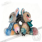 Two vibrant, furry plush toys sit back to back. Each features oversized grinning mouths with sharp red teeth, large round eyes, and blue bows—capturing the whimsical essence of the Victorian McGee's Autism Acceptance Cheshire Cat Plush Stuffed Animal. Their fur is patchworked in soft pastel hues of blue, pink, yellow, and gray, making them a playful homage to Alice in Wonderland characters and a touching symbol of autism acceptance.