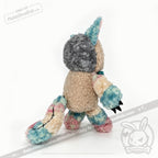 Victorian McGee's Autism Acceptance Cheshire Cat - Plush Stuffed Animal is pictured from the back against a white background, displaying pastel blue, pink, and beige fur; a grey head; unicorn horn; long tail with black stitches; and black claws. Perfect for fans of Alice in Wonderland and autism acceptance alike.