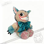 A vibrant Victorian McGee's Autism Acceptance Cheshire Cat plush stuffed animal—with multicolored fur, oversized pointy ears, a broad toothy grin, expressive large eyes, and a teal bow around its neck—rests on a white background, joyfully celebrating autism acceptance.