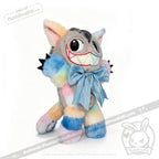 Standing on all fours against a white background, the Victorian McGee's Autism Acceptance Cheshire Cat Plush Stuffed Animal features vibrant rainbow-colored fur, a big embroidered toothy grin, and a large blue bow around its neck—an enchanting plush inspired by Alice in Wonderland, perfect for spreading joy and autism acceptance.