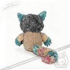A Victorian McGee's Autism Acceptance Cheshire Cat plush stuffed animal—with a round beige body, blue ears, multicolored arms, and a long fluffy tail—is shown facing away from the camera on a white background. This Alice in Wonderland-inspired plush is designed to promote autism acceptance.